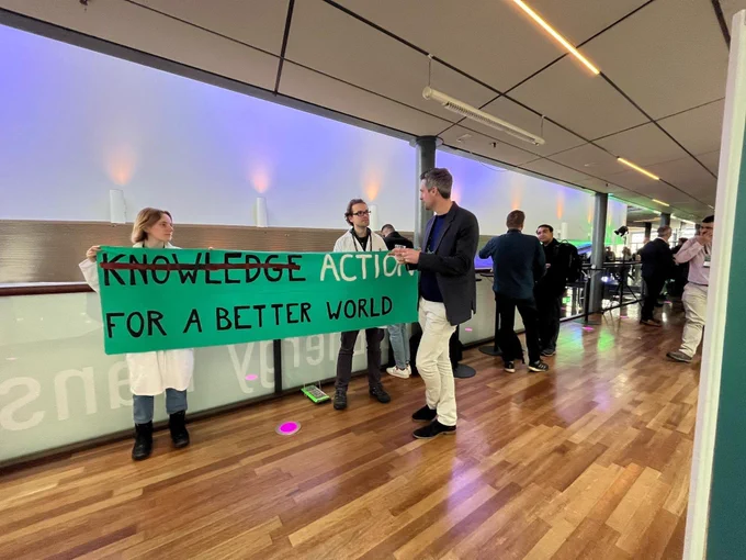 Two scientists in lab coats holding a banner reading "<strikethrough>Knowledge</strikethrough> Action for a better world" talk to a participant at the NTNU Energy Transition Conference