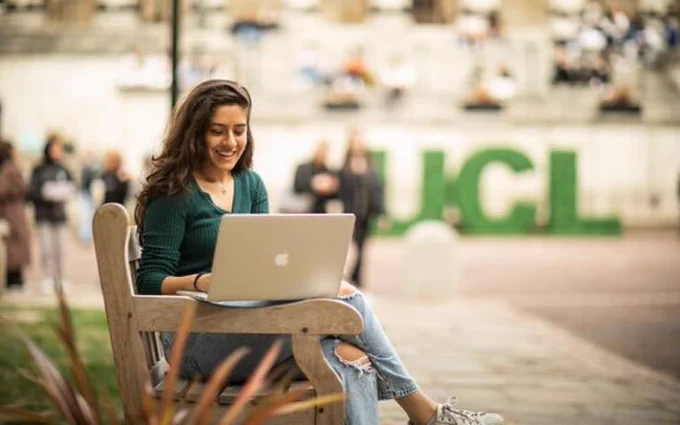 Photo of a smiling woman working on a laptop on a bench outside UCL. 