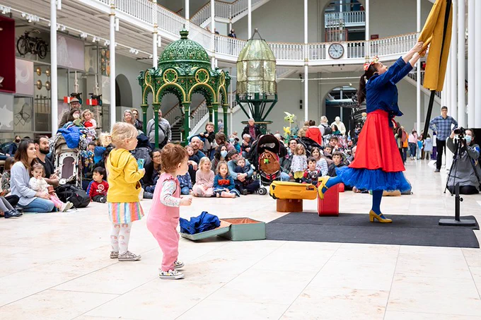 Decorative image from Family Encounters. A performer in a long red and blue skirt setting up a yello peice of fabric above their head. A group of young children and parents sit on the ground of the main museum hall watching in the background. In the foreground two toddlers are standing and watching the performer. Photo credit Ruth Armstrong. 