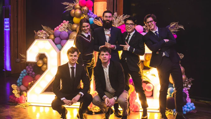 A group of 6 students posing together in front of a large display of light-up numbers and colourful balloons. One of the students is holding an award.