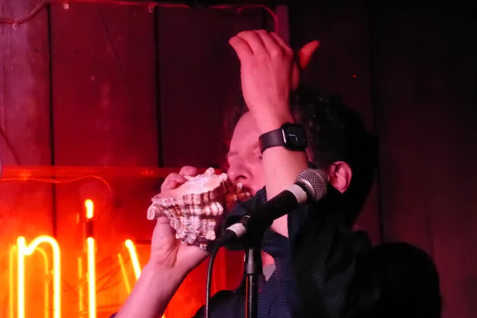 Steve looks nowhere, eyes closed in concentration as he plays a conch like a trumpet
