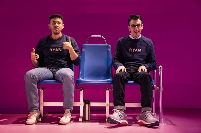 Dickie Hearts and Ryan J. Haddad sit on blue and silver metal bus benches with a pink projection behind them. Ryan is a Lebanese American man with short black hair, brown eyes, and round, tortoiseshell glasses. Ryan wears a navy blue long-sleeve sweatshirt with the word “RYAN” printed in white across his chest and a white button-down shirt underneath it. He wears blue jeans and grey and white Nike sneakers with red and black soles. Ryan looks straight out to the audience and smiles with his hands resting on his chest. Dickie Hearts has olive skin and dark chocolate-colored curly hair. He also wears a navy blue long-sleeve sweatshirt with the words “RYAN” printed in white across his chest. He wears light blue jeans and white sneakers with rainbow soles. Dickie two seats down from Ryan with his legs spread out. He communicates in ASL with his eyes looking up towards his left.