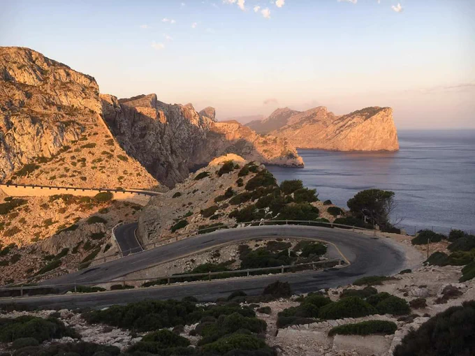 A winding road runs along the coast of Majorca. A dry landscape with scattered bushes and steep Cliffs down to the sea. 