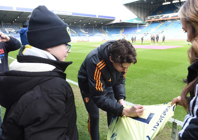 Brenden Aaronson signing the t-shirt Leeds FC gifted to Alfie. Alfie is watching Brenden sign the t-shirt. They are on the football pitch in the stadium grounds.