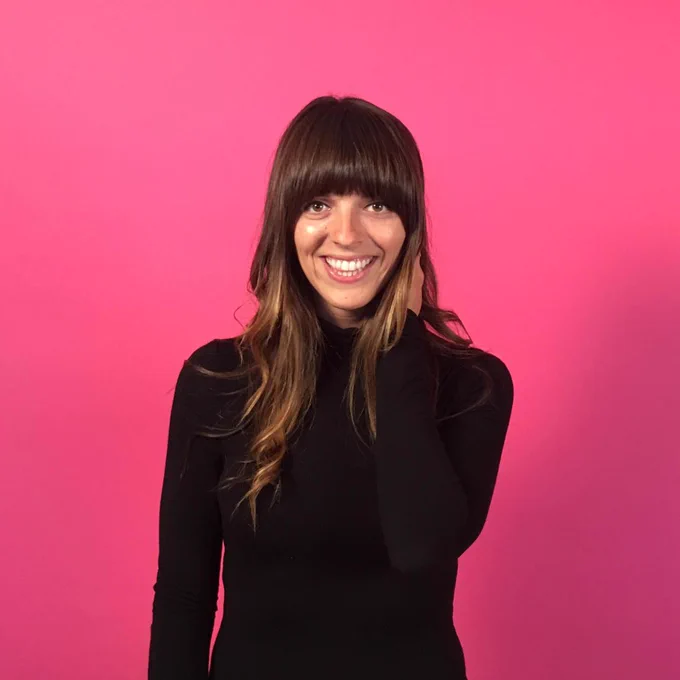 Headshot of a woman with long brown hair and blunt blangs smiles and looks towards the camera. She wears a black turtleneck and pants and stands in front of a pink background.