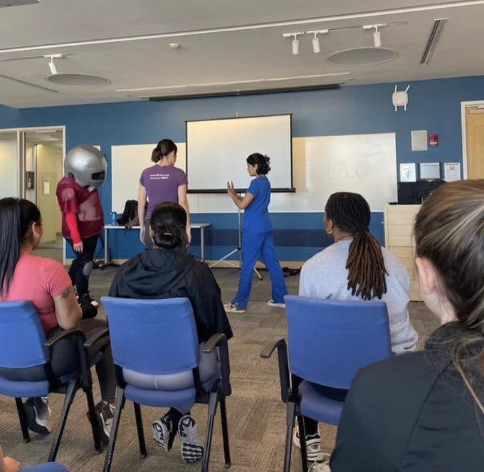 A student standing with stop sign hands up in a self-defense scenario, prepared to use physical skills. A suited instructor wearing their helmet stands in front of the student ready to move in. Another instructor stands off to the side/between them to help the student. Other students are seated and watching.