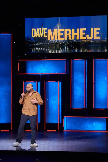 Dave Merheje on stage with a microphone under a sign that says his name in yellow 