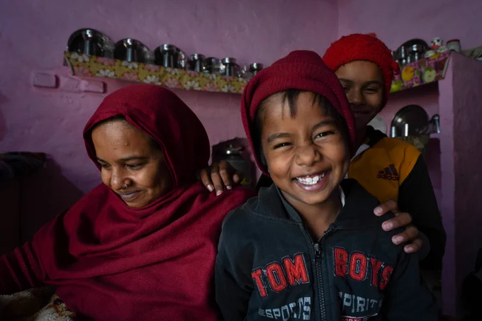 A woman wearing a maroon headscarf smiles shyly with two children, one is scrunching his nose in a cheeky grin