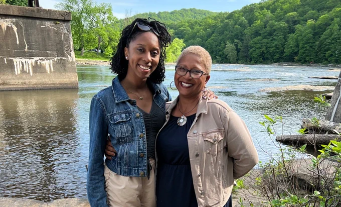 Lauren and Laurita stand together in front of a river with lush green hills in the background