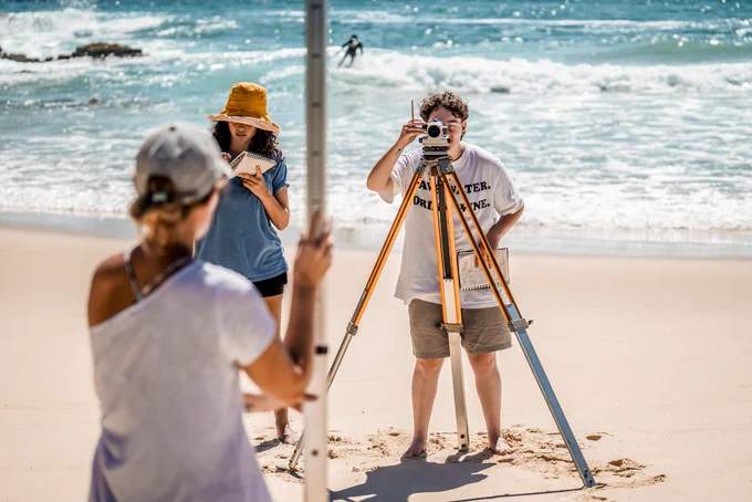 Two students are working on a science study on the sandy shores of a beach. One is writing on her notebook and the other is looking down the lens of equipments resting on a tall tripod. They are both wearing casual clothes and no shoes. 
