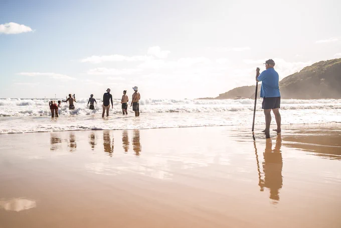 A man in black shorts and a blue shirt is holding a long measuring ruler as he watches 6 people in knee deep water at the beach on a sunny day. They have various equipment to help study coastal processes. 