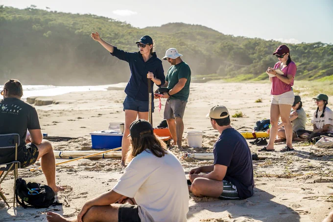 A group of students dressed in casual sun protective clothes are sitting on the sandy shores of a beach on a sunny day as they listen to a scientist dressed in navy shorts, top and cap as she points out into the water with her hand. 