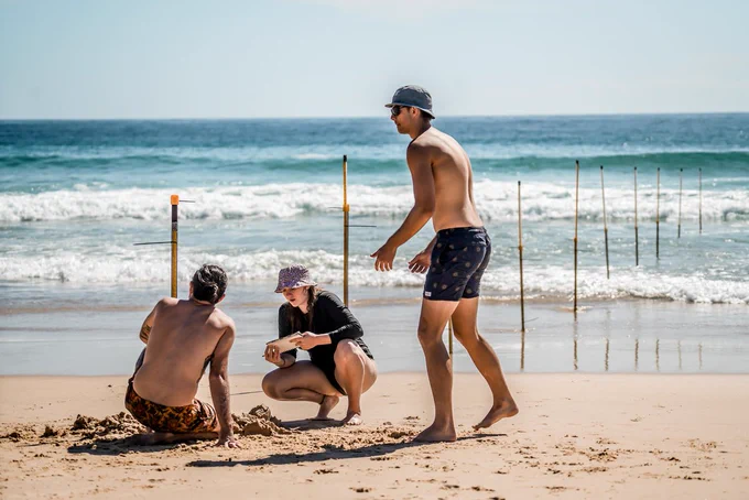 Three students wearing swimmers are gathered on the sand as tall markers are set in a line in the ocean waters behind them. 