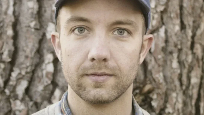 Close-up of Jon Klassen looking at the camera. He is in front of a tree trunk.