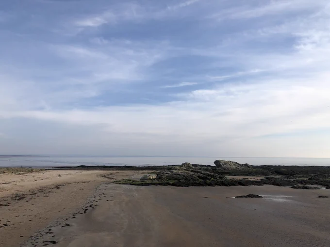 Photograph of a beach with lots of big rocks and sand looking as though the tide has only recently gone out. The sea is very pale blue in the distance and almost blends in with the sky which is a pale blue/grey with streaks of clouds.