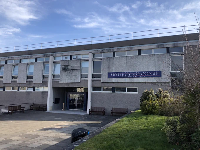 Photograph of concrete building with a very square design and lots of windows. There’s a courtyard in front and some grass and bushes on the right, on a slope. The sky is vert blue with a few clouds.