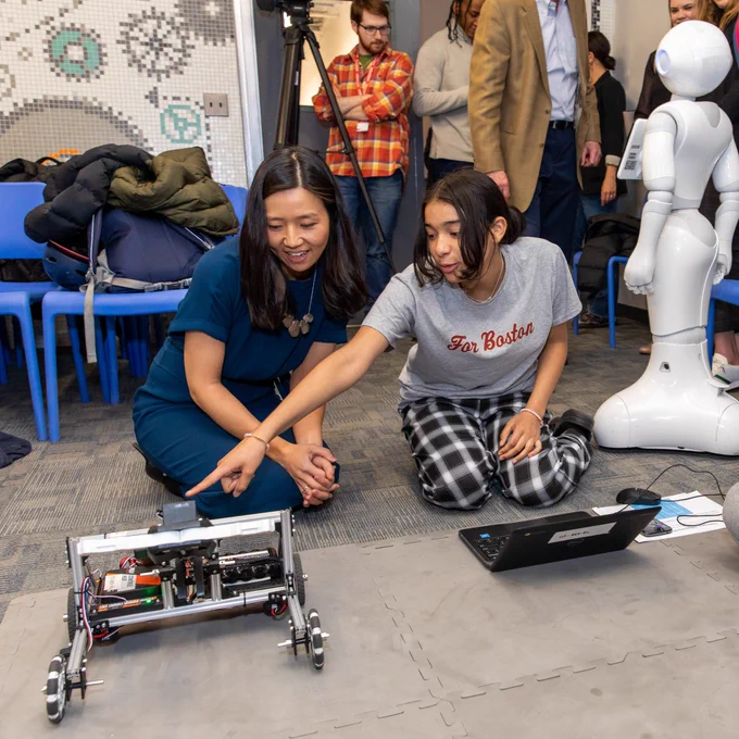 Two young community members and Mayor Wu sit on the floor together. There is a laptop sitting in the middle of them. One person points to a robot, showing the Mayor something. There is a person-like robot standing off to the side, with other people in the background.