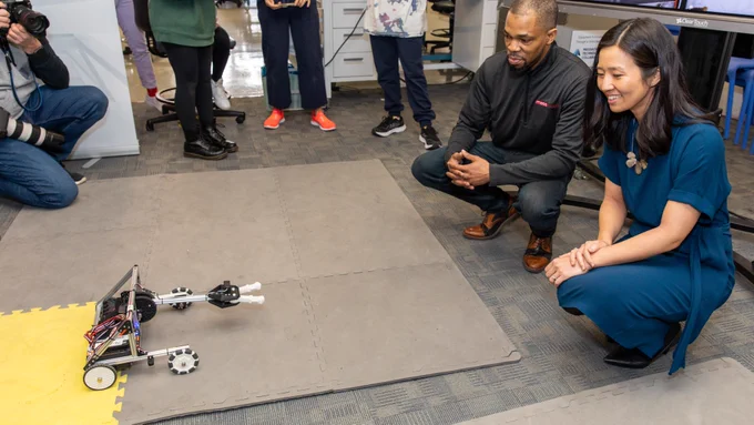 A person from MassRobotics crouches down with Mayor Wu as they look at a robot sitting on a padded mat. There are a few people standing in the background.