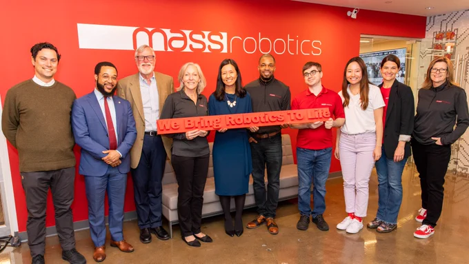 MassRobotics leaders, Chief Idowu of the City of Boston, and Mayor Wu come together for a photo. Four people, including the Mayor, hold a red sign that reads, "We Bring Robotics To Life". They all stand in front of a red wall featuring the "massrobotics" logo.