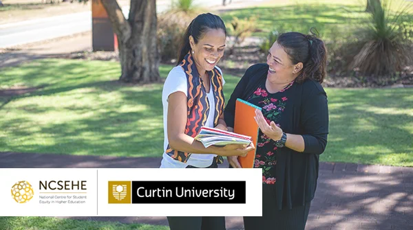 Two adult women, talking and smiling, holding books, whilst standing under a tree in a park-like are.