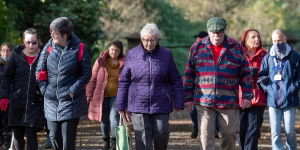 A large group of people of all ages and abilities in outdoor clothing enjoy a walk during a sunny day along a country path surrounded by woodland.