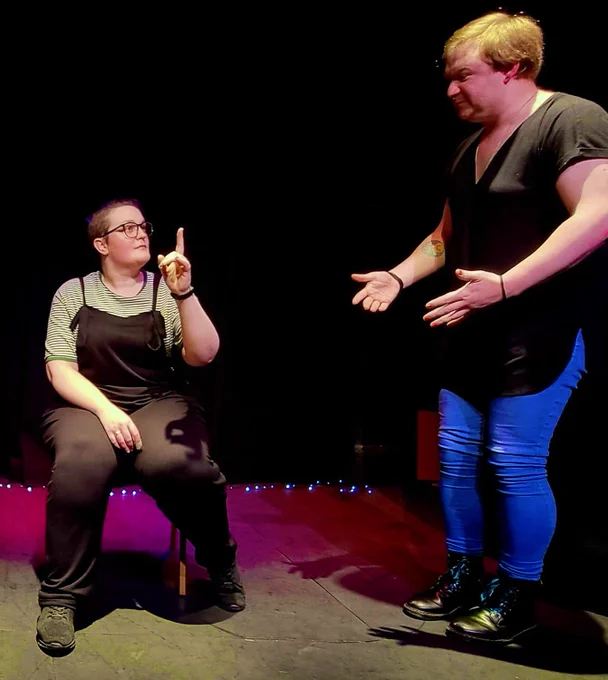 Two people onstage, one is sat down gesturing for silence, the other reaches out towards them exasperated