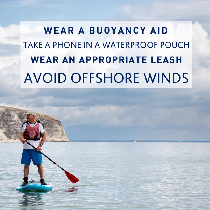 A man is in the sea, wearing a lifejacket, stands on a blue paddleboard.