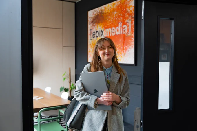 Marty from Team Epix standing in the doorway to a meeting room, carrying her laptop under her arm. There is a large Epix Media mural on the wall in the meeting room and a meeting room table and chairs.