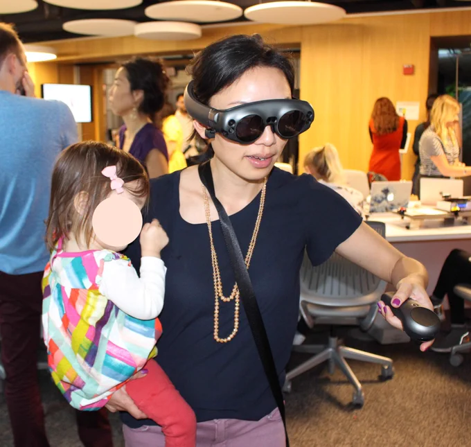 Asian woman wearing a navy shirt and 2-strand peach necklace and a Magic Leap headset over her eyes and across the back of her head. The headset is dark gray metal and plastic and looks like dark goggles. She has a strap across shoulder holding the other hardware. She is playing a game while holding a controller in left hand and a young child in a colorful dress and red pants on her right hip and in right hand. People are in the background talking in a meeting space with tables and office chairs.