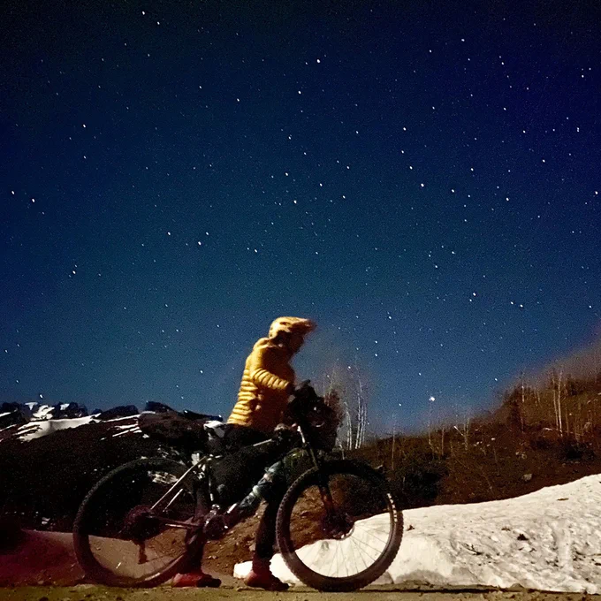 Cat Jaffe, of @houseofpod, in a yellow jacket and yellow helmet, walks her bike under a starry sky in the desert. Photo by Cat Jaffe.