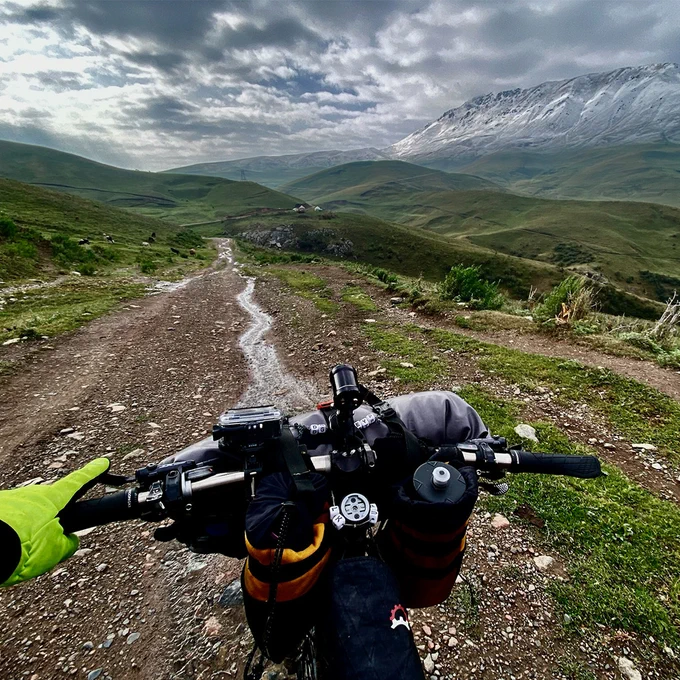 Photo taken by Cat Jaffe, of @houseofpod, from her bike. We can see her left hand holding the handlebar. Ahead of her, a dirt road faces a landscape of green hills under a cloudy sky. On the right, a chain of snowy mountains. 