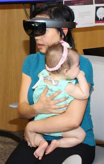 Asian American woman sitting in an office chair. She is wearing a teal short sleeve shirt and black pants. Covering her eyes to the top of her nose is a black and gray HoloLens headset, which wraps around the back of her head. She holds an infant baby in her crossed arms. The baby's back is pictured,  wearing an aqua colored onesie with a silver bow on the back and a pink stretchy headband across the back of her head. The baby is chewing her right fist.