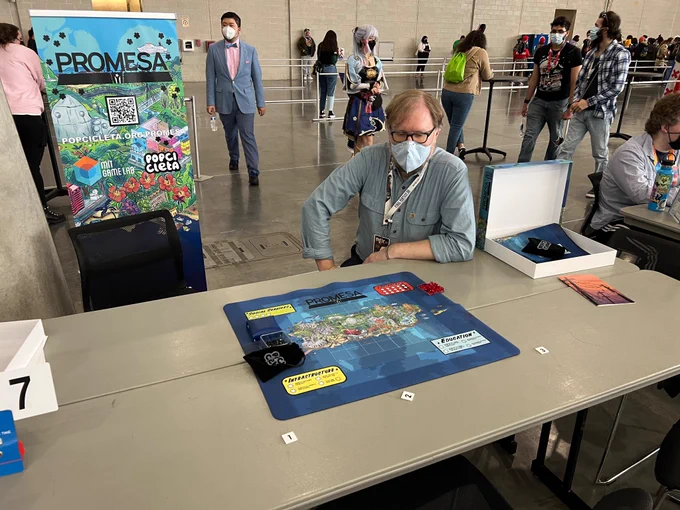 Promesa board (a neoprene May with an illustrated map of Puerto Rico) set up on a table with Mikael staring intently at it. A banner is beside him with artwork from the game and a QR code for the website popcicleta.org/promesa