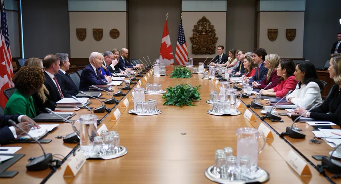 Prime Minister Justin Trudeau and President Joe Biden are sitting across from each other at a large table. Cabinet secretaries are sitting beside the President, and Cabinet ministers are sitting beside the Prime Minister. Other officials and staff are also sitting at the table. Canadian and American flags are in the background.