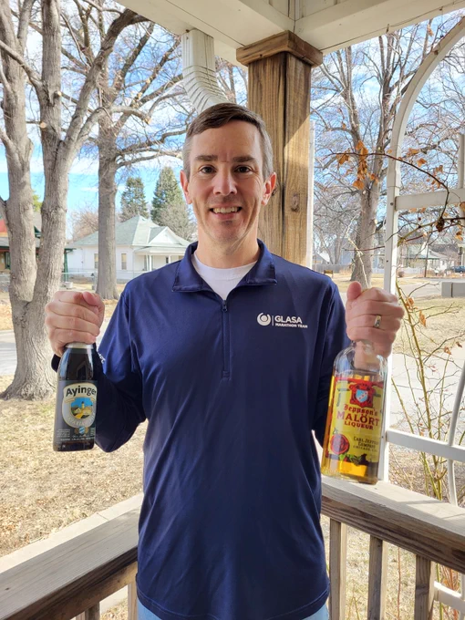 A man smiles wearing wearing a quarter zip with the Great Lakes Adaptive Sports Association (GLASA) logo on his chest pocket while holding a bottle of Jeppson's Malört in one hand and a bottle of Ayinger Bavarian Pils in the other.
