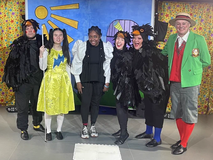 Actors & BSL interpreter stand in front of the set. The set is a park scene with green grass, blue sky, yellow sun & purple observatory. Either side are yellow curtains with coloured stars. A big white jigsaw square is on the floor in front of them. Left to right - Steve George is a white actor dressed as a black bird with a black bird head with orange beak & black feathers with a black feather cape, black trousers & black shoes, next is Jasmin Thien. She’s Chinese, slim & has long black hair in bunches, wearing a yellow dress with a blue flash across her chest. She has her long white cane in her right hand. Next is BSL interpreter Debbie who’s black with long braids wearing a white shirt with puff sleeves & black sleeveless dress over the top. Next is Angela who is white & Chloe who is of mixed heritage both in black bird costumes. Finally is Chris Biddle who’s white with a green blazer, red shirts, straw boater hats, grey shorts & long red socks. Everyone is smiling at the camera.