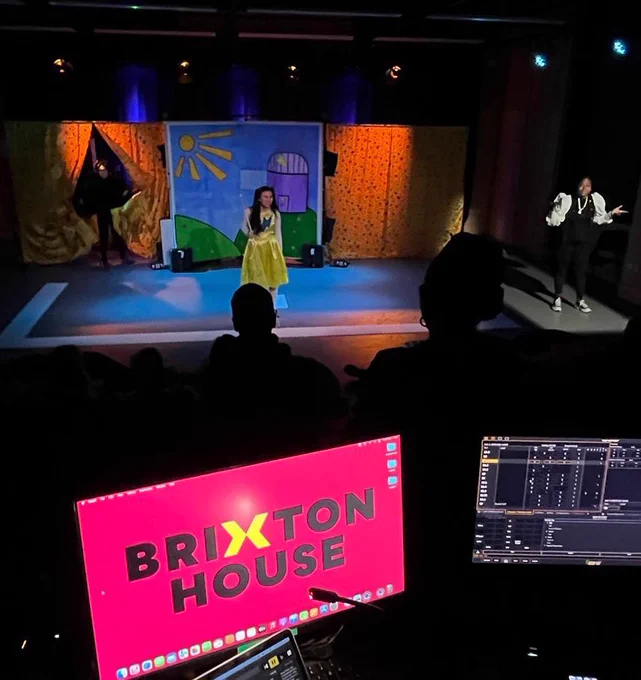 2 screens at angles at the bottom on the photo - On the left is a pink screen with ‘Brixton House’ in large black letters with a yellow x. Right is a tech screen for sound queues. There are lights above the stage. Silhouettes of the audience can be seen. The set is a park scene with green grass, flowers, blue sky, yellow sun & purple observatory. Either side are yellow curtains with red, blue & green stars. Actress Angela Eyton who’s white enters the curtain on the left in her black bird costume with a big orange beak on her head & black feathers on her cape. Centre stage is Jasmin Thien. She’s Chinese, slim & has long black hair in two bunches, wearing a pale-yellow  dress with a blue flash across her chest. She stands in front of the set holding her long white cane in her right hand on a white square with jigsaw edges. On the right in the spotlight is Deborah who is a black woman signing BSL. She is wearing a puff white shirt with a black sleeveless dress over the top & trainers