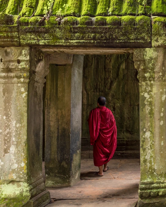 A colour image of some columns in a Cambodian Temple and a buddhist monk wearing red robes is walking away from the camera and into the temple.