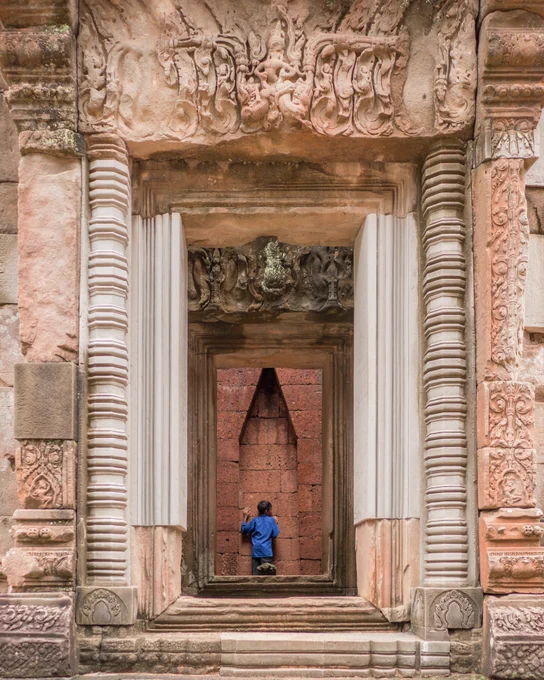 a photograph of an intricate doorway in a buddhist temple in Cambodia that leads through to a well lit room, probably due to there being no roof, and a wall at the end. There is young boy in a blue t-shirt leaning on the wall almost as though he's listening to the walls, the history of his ancestors. 