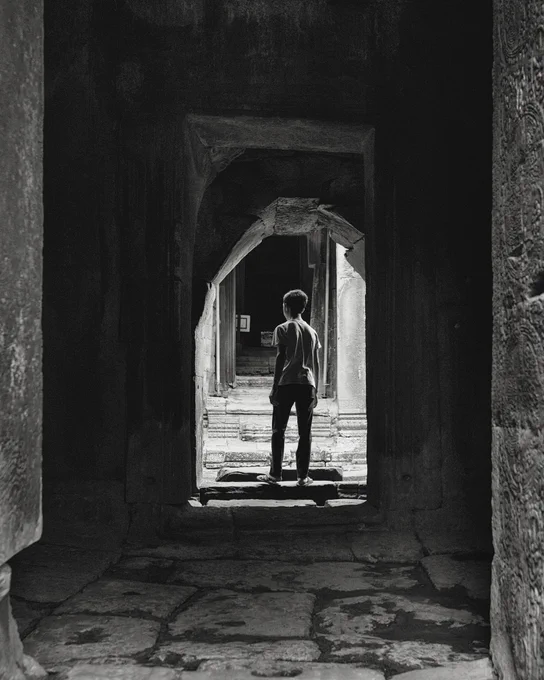 A black and white photo of a boy standing in a doorway, facing away from the camera, looking like he's making a decision as to which direction he is about to go. 