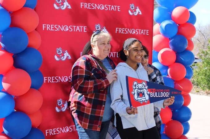 Student with her family in front of a Fresno state backdrop and red and blue balloons