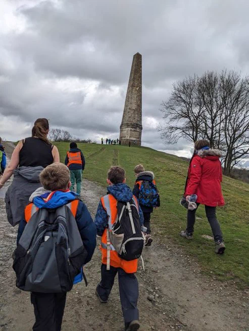 5 young people & 2 adults wearing back packs walking away from the camera towards a column monument.