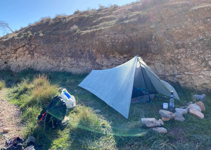 Tent on grassy creekbed sheltered by red earth creek bank, sun streaming down from a blue sky