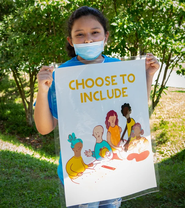 A young student holds a sign that says "Choose to Include" with an illustration of five diverse people below the text.