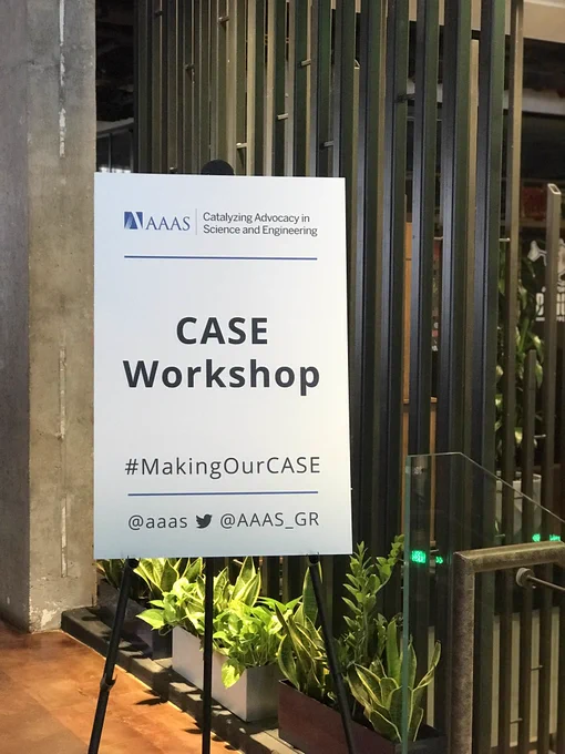 Sign promoting the AAAS CASE Workshop and it's social media handles, with decorative plants in the background.