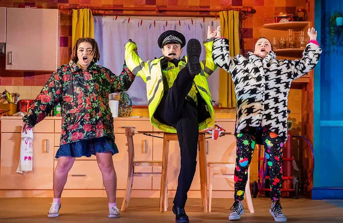 Three performers on stage holding hands leaning backwards. The performer in the middle is dressed as a policeman with one leg up in front of him. There is a kitchen set behind them 