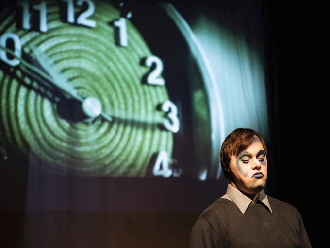 A performer wearing abstract makeup in shades of blue, look sad and 'blue'. They stand in front of a large projection of a green clock. 