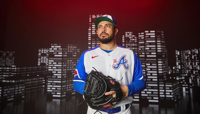 Travis d'Arnaud poses in the Braves' City Connect Uniforms while wearing a catcher's mitt. The uniforms consist of a blue hat with a white panel, a white jersey with blue sleeves with "The A" on the right chest, and the player's number on the lower left side of the jersey. The pants are white. 