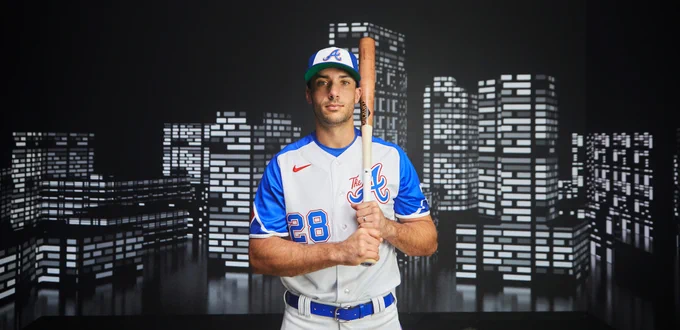 Matt Olson poses in the Braves' City Connect Uniforms while holding a bat on his left shoulder. The uniforms consist of a blue hat with a white panel, a white jersey with blue sleeves with "The A" on the right chest, and the player's number on the lower left side of the jersey. The pants are white. 