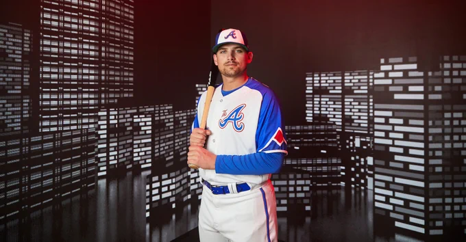 Austin Riley poses in the Braves' City Connect Uniforms while holding a bat on his right shoulder. The uniforms consist of a blue hat with a white panel, a white jersey with blue sleeves with "The A" on the right chest, and the player's number on the lower left side of the jersey. The pants are white. 